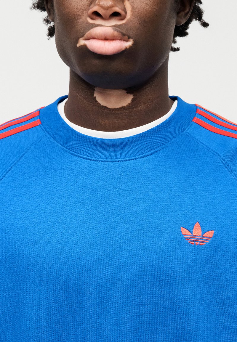 Sweat-shirt bleu en coton, avec un col rond, des accents rouges à trois bandes sur les épaules, et un petit logo brodé en rouge.