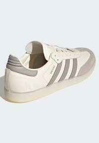 Zapatilla blanca de cuero con acentos en gris, diseño de tres franjas, capucha de ante en la punta y lengua acolchada. Presenta un logotipo en relieve en el talón.