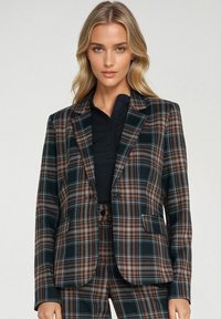 Marynarka w kratę w ciemnozielonych, pomarańczowych i brązowych odcieniach. Posiada dwie przednie kieszenie, wycięty kołnierz, zapięcie na pojedynczy guzik oraz miękką teksturę tkaniny.