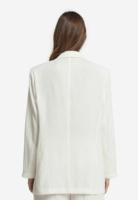 Kvinna med långt brunt hår som bär en lös vit kavaj, stående med ryggen mot kameran framför en vanlig vit bakgrund.