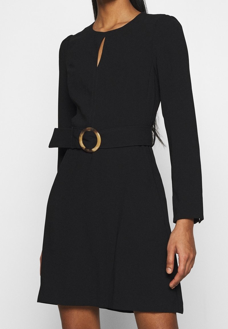 Černé šaty s kulatým výstřihem, dlouhými rukávy, předním klíčovým detailem a páskem v pase s kruhovou, želvovinovou sponou.