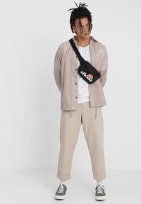 Light pink, shiny jacket over a white t-shirt, paired with beige, tapered pants and green sneakers. Black crossbody bag has logo accent.