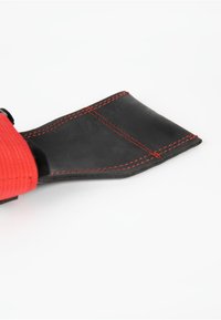 Base en caoutchouc noir avec couture rouge, attachée à une sangle rouge texturée. Le design est plat avec une forme rectangulaire et des bords nets.