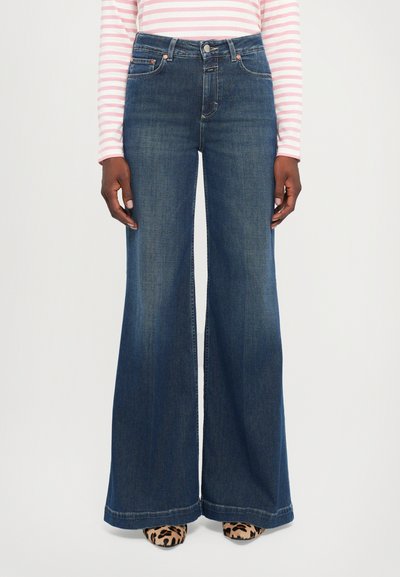 Schwarz gewaschene, ausgestellte Jeans mit vorderen Taschen und Gürtelschlaufen. Sie haben eine hohe Taille und ein weites Bein-Design, kombiniert mit Schuhen im Tiermuster.