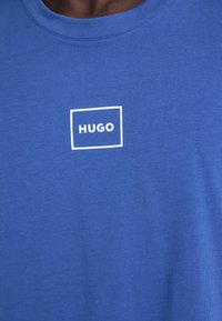 Modrá bavlnená tričko má okrúhly výstrih. Na prednej strane je umiestnené biele obdĺžnikové logo s textom "HUGO".