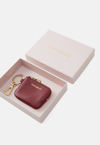 Burgundy leather coin pouch with gold hardware, zip closure, and keychain loop, presented in a pink box with gold brand text.