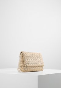 Beżowa pikowana kopertówka z aksamitu z trójkątnym wzorem, klapą na zakładkę i metalowym akcentem. Gładka faktura, zorganizowany kształt, minimalistyczny design.