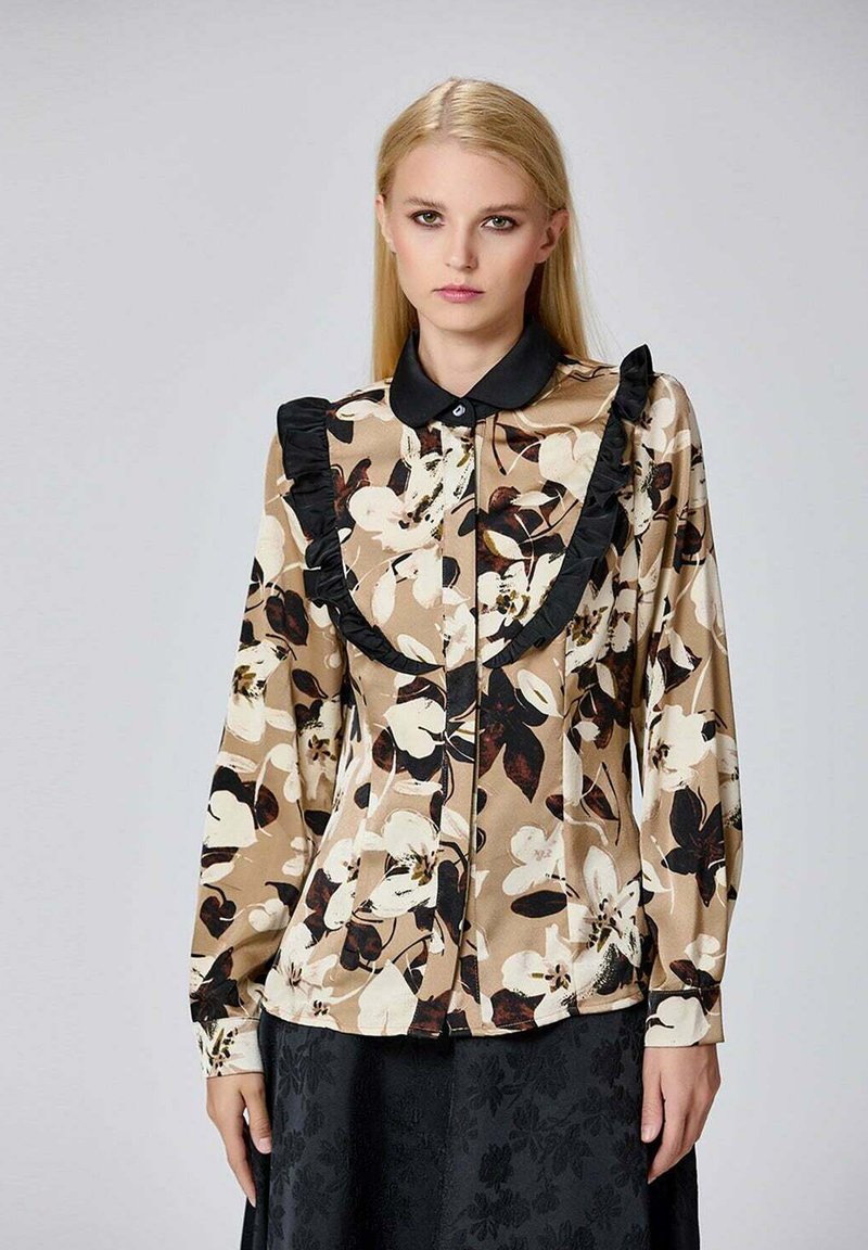 Blouse florale beige, avec un trim à volants noir le long du col et une fermeture à boutons. Manches longues, tissu lisse et motif floral audacieux.