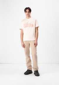 Helles rosa T-Shirt mit kurzen Ärmeln und orangefarbenem Lacoste-Logo, kombiniert mit hellbeigen Hosen und schwarzen strukturierten Sneakern. Steht vor einem weißen Hintergrund.