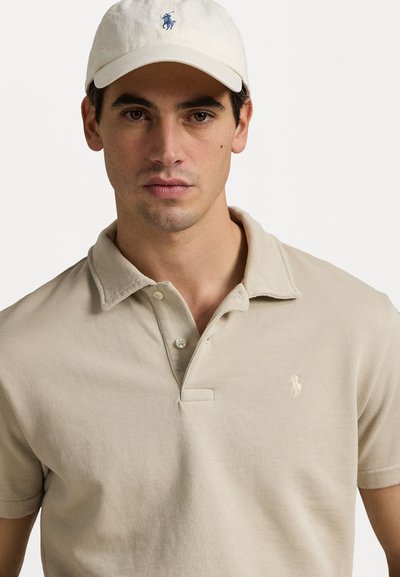 Polo beige avec un col classique, deux boutons et un logo brodé. Porté avec une casquette de couleur claire. Texture lisse.