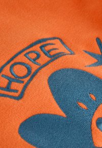 Oranž kangas, millel on sinine tikandiga disain, mis ütleb "LOOTUS", lillekuju ja väikeste leheaktsentidega. Pehme tekstuur, puhas viimistlus.
