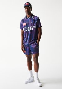 Αθλητική μπλούζα με μοτίβο σε μωβ και μπλε χρώμα με λογότυπο "ORBR", συνδυασμένη με αντίστοιχα σορτς. Λευκές κάλτσες και αθλητικά παπούτσια συμπληρώνουν την εμφάνιση. Καπέλο με το λογότυπο.