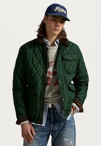 Homme portant une veste matelassée vert foncé, un sweatshirt gris, une chemise rayée, un jean bleu et une casquette bleue avec le texte "Ralph", se tenant avec une main dans sa poche.