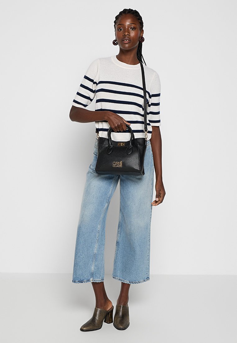 Bolso de mano de cuero negro con logotipo en relieve, forma estructurada, doble asa superior y correa de hombro desmontable. El modelo lleva un suéter a rayas y jeans de pierna ancha.