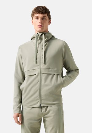 Jonge man met een lichtgrijze zip-up hoodie met trekkoorden en bijpassende broek, staand met één hand in de zak tegen een effen achtergrond.