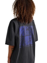 Osoba z kręconymi włosami ubrana w luźną czarną koszulkę z niebieskim abstrakcyjnym wzorem siatki na plecach, stojąca na białym tle.