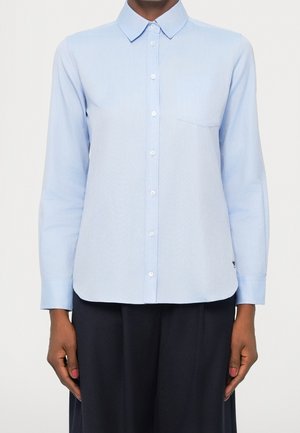 Chemise bleu clair à manches longues avec boutons, col et poche poitrine, portée sortie sur un pantalon foncé.
