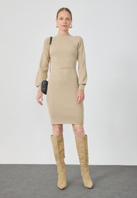 Beige ribbet strikkjole med høj halsudskæring, lange pufærmer, en tætsiddende talje med et gyldent accent, parret med brune knæhøje støvler.