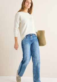 Helles beiges Rippstrickpullover, blaue gerade Jeans und graue Sneakers. Hält eine olivgrüne Puffertasche. Neutrale Hintergrund.