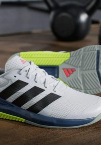 Zapatillas deportivas blancas con rayas negras, detalles grises y una parte superior texturizada. Presentan una suela colorida con detalles en verde y rojo.