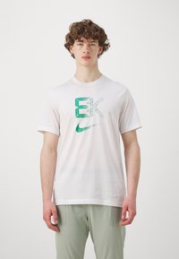 Hvit t-skjorte med kort erme og en teksturert grønn og svart grafikk som har bokstavene "EK" og en grønn Nike-logo. Laget av bomull.