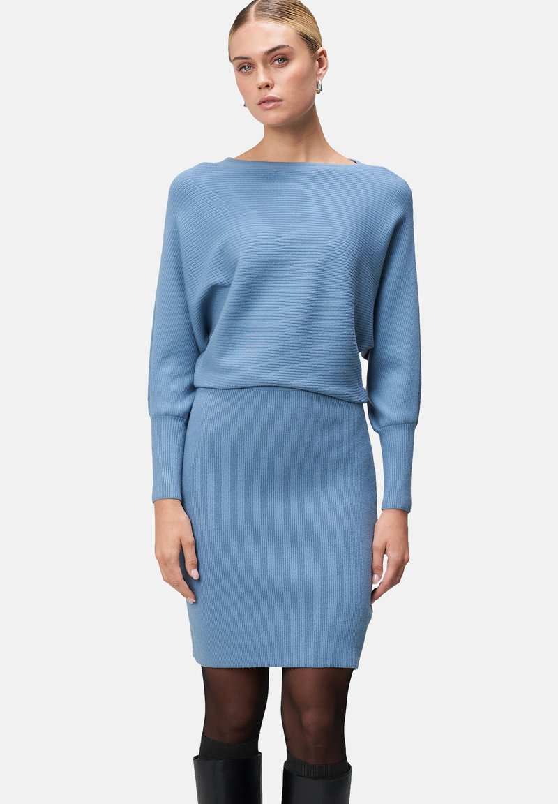 Robe pull en tricot côtelé bleu clair, avec une coupe décontractée, des manches longues et un col bateau, associée à des bas noirs et des bottes montantes.