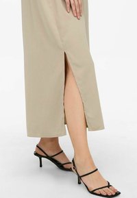 Beige midi-jurk met zijsplit, gemaakt van gladde stof, gecombineerd met zwarte sandalen met smalle hak en bandjes.