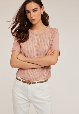 Top scurt de culoare roz deschis, cu mâneci scurte, având un design cu paiete pe față, gât rotund, asortat cu pantaloni albi și o curea de piele maro cu un fermoar decorativ.