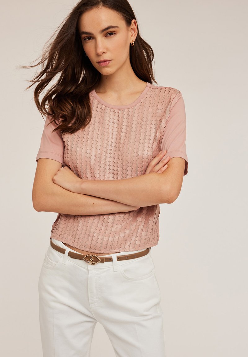 Top scurt de culoare roz deschis, cu mâneci scurte, având un design cu paiete pe față, gât rotund, asortat cu pantaloni albi și o curea de piele maro cu un fermoar decorativ.