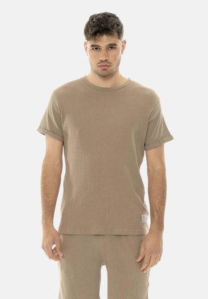 Jeune homme aux cheveux courts et foncés portant un t-shirt côtelé beige à manches courtes et un pantalon assorti, debout devant un fond uni.