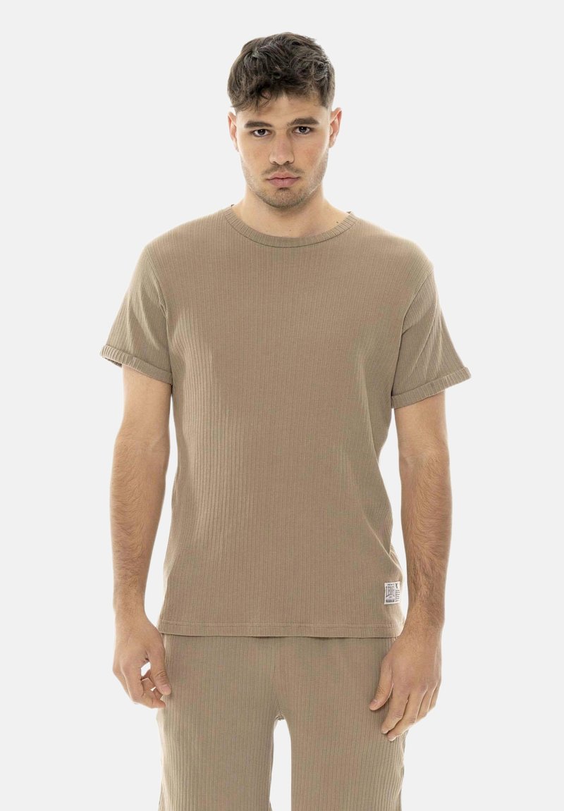 Jeune homme aux cheveux courts et foncés portant un t-shirt côtelé beige à manches courtes et un pantalon assorti, debout devant un fond uni.
