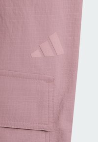 Tissu violet clair avec une poche rectangulaire cousue et un logo Adidas en relief dans une tonalité violette assortie.