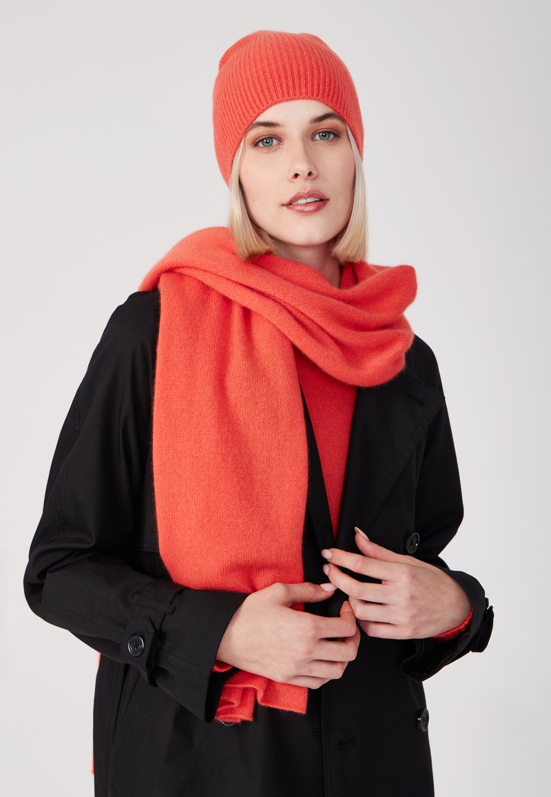 Cappello e sciarpa in lana corallo abbinati a un cappotto nero. La sciarpa cade morbida, mettendo in mostra una texture soffice e un colore vivace.