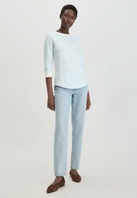 Sininen ja valkoinen raidallinen pitkähihainen paita, yhdistettynä vaaleansinisiin farkkuihin ja ruskeisiin kärkitossuihin, seisoo neutraalia taustaa vasten.