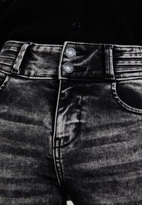 Jean en denim noir avec une finition usée, présentant deux fermetures à bouton argentées, des poches visibles et un tissu texturé.