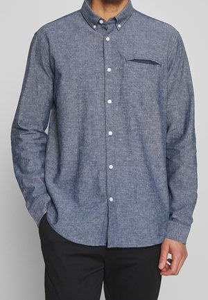 Homme portant une chemise chambray bleue à manches longues, boutonnée, avec col plié et poche passepoilée, associée à un pantalon noir.