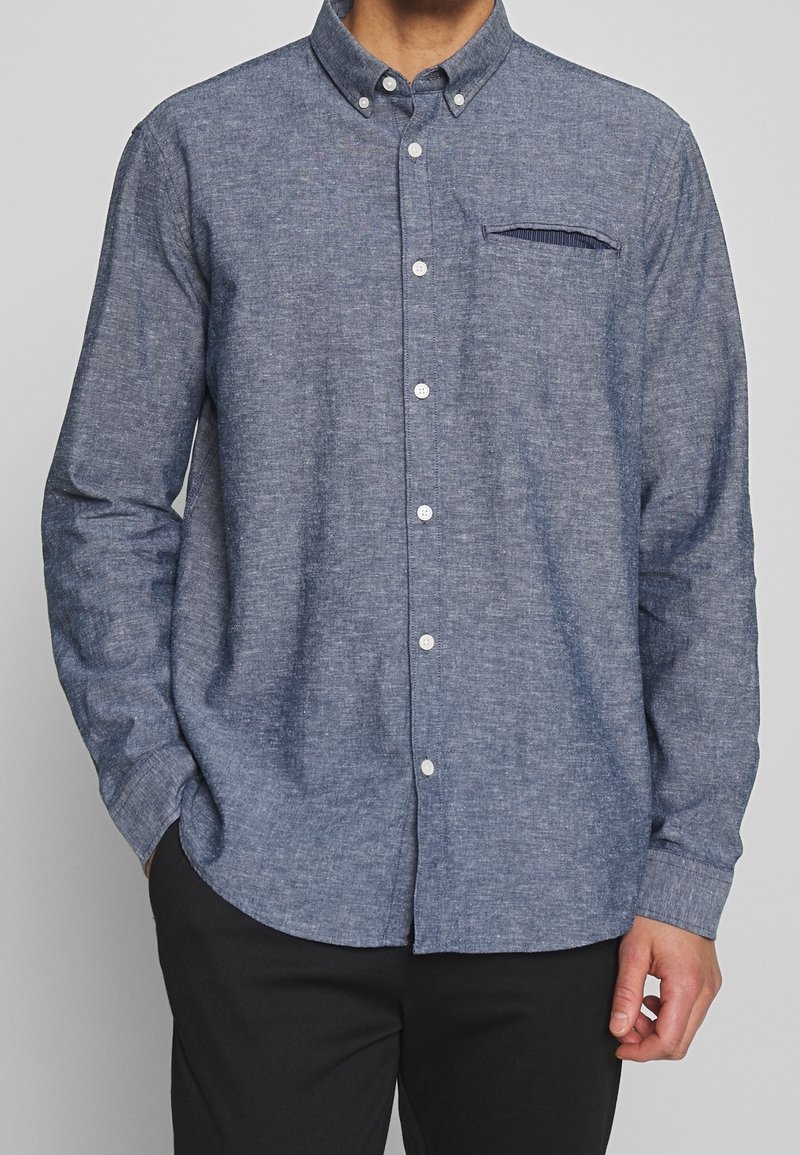 Homme portant une chemise chambray bleue à manches longues, boutonnée, avec col plié et poche passepoilée, associée à un pantalon noir.