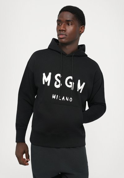 Giovane uomo che indossa una felpa nera MSGM Milano con tasca frontale, in piedi contro uno sfondo chiaro uniforme, mentre guarda di lato.