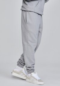 Graue Sweatpants mit elastischen Bündchen, glatter Textur und Seitentaschen, kombiniert mit weißen und grauen Sneakers mit Wellenmuster und Schnürsenkeln.