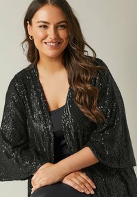 Veste kimono noire en sequins avec un devant ouvert, tissu texturé et manches larges, superposée à un top noir au surface lisse et brillante.