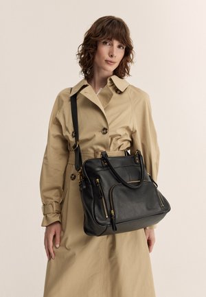 Bolso de mano de cuero negro con cremalleras y acentos dorados, que cuenta con dos asas y una correa de hombro desmontable, colocado sobre un fondo neutral.