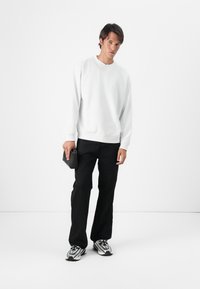 Sweatshirt branco com decote redondo, emparelhado com calças pretas de perna reta. A pessoa segura uma bolsa de mão preta texturizada e usa ténis prateados.