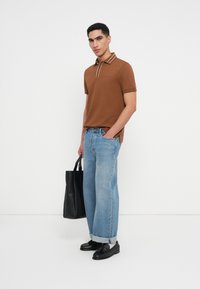 Brūna polo kreklu ar baltu apdari, kombinēts ar gaiši ziliem platām biksēm. Modelis tur melnu somu un valkā melnas kurpes ar zemu papēdi.