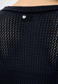 Gros plan sur le dos d'un pull en maille noire tricotée laissant apparaître la peau en dessous et une petite étiquette ronde en argent près du col.