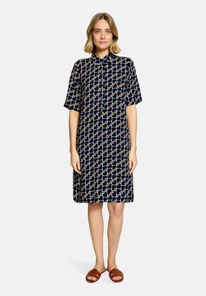 Kvinde med kort blondt hår iført en kortærmet, knælængde navy kjole med et beige geometrisk mønster og brune slidesandaler.