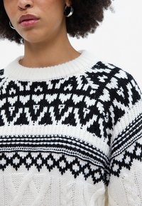 Mustra musträkistatud must-valge nõelumine, millel on soonikuga ümar kaelus, tekstuuriga kaablitooted ja geomeetrilised mustrid kogu keha ulatuses.