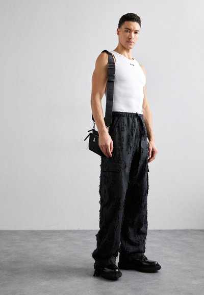 Pantalon cargo noir avec une texture effilochée, porté avec un débardeur blanc ajusté. Un sac à bandoulière noir pend d'une sangle sur l'épaule. Chaussures noires.