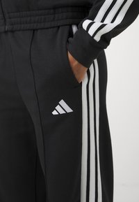 Svarta sweatpants med tre vita ränder längs sidorna och en liten vit Adidas-logotyp nära fickan. Gjorda av mjukt material.