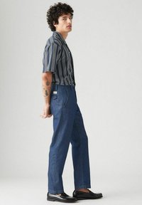 Nuori mies, jolla on kiharat hiukset, seisoo sivuttain. Hänellä on päällään raidallinen lyhythihainen paita, siniset farkut ja mustat loaferit. Hänen kyynärvarren tatuointinsa ovat näkyvissä.