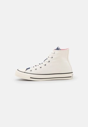 Zapatilla blanca de lona de caña alta con cordones blancos, suela de goma y detalle de una franja negra, vista de lado sobre un fondo blanco.